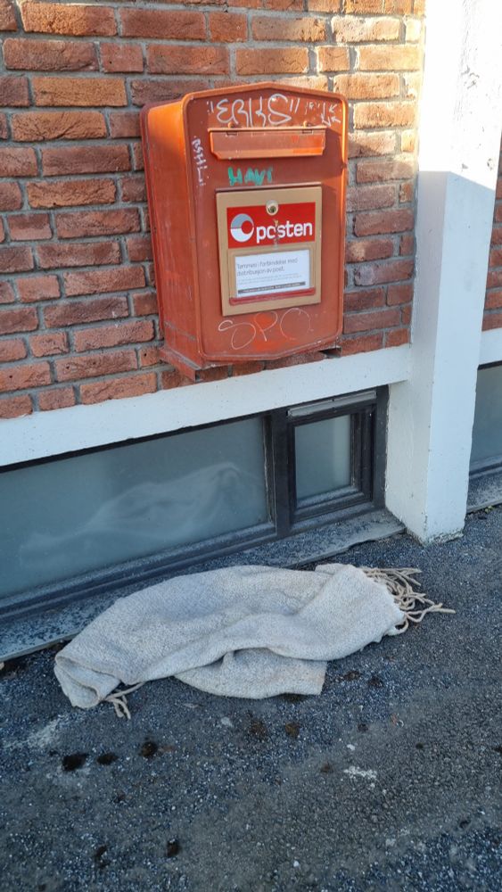 Et pledd eller et veldig stort skjerf som ligger på bakken under en Posten postkasse. Refererer til et mye omtalt VELDIG stort skjerf Lenny Kravitz ble avbildet med en gang. Som ble en meme.