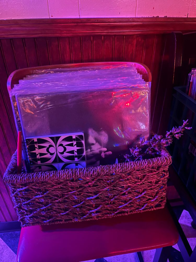 Photo of a woven crate on a chair containing records (a Maki Asakawa compilation being visible,) a notebook with a marker, and a sprig of sage flowers