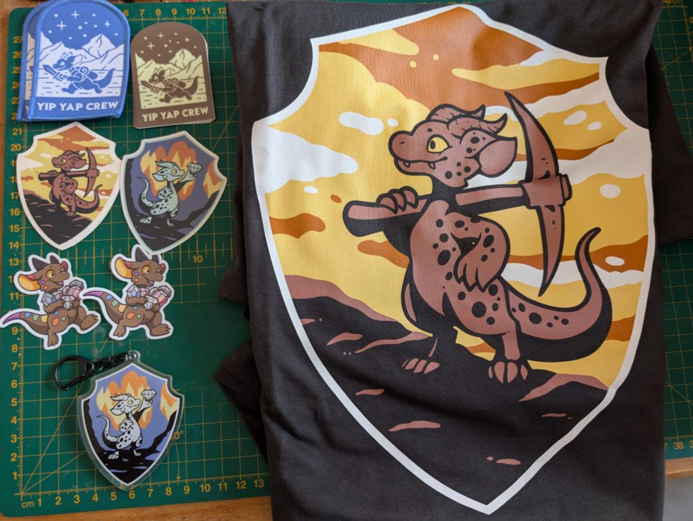 Photo of a large t-shirt with a kobold brandishing a pickaxe with a number of mining themed kobold patches, stickers and charms