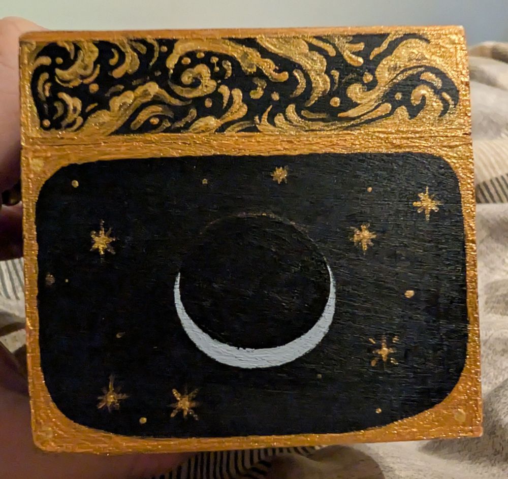 Photo of a side of a small wooden jewellery box painted with gold filigree, stars and silver moon on a black background