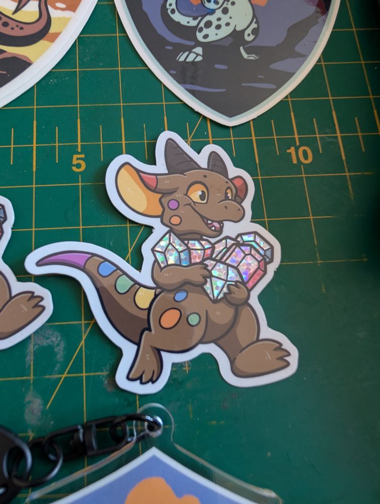 Close up of a kobold sticker, their spots are in the pride flag colours and they are carrying glittering, trans pride coloured gemstones 
