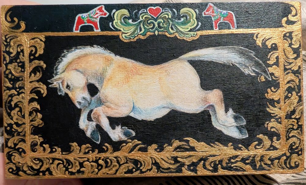 Photo of the top of a small wooden box painted with gold filigree, dala horse figurines and centrally a jumping fjord horse