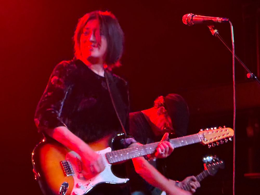 Mid range closeup of Miki Berenyi playing guitar and smiling as she’s engulfed in a red backlight. 