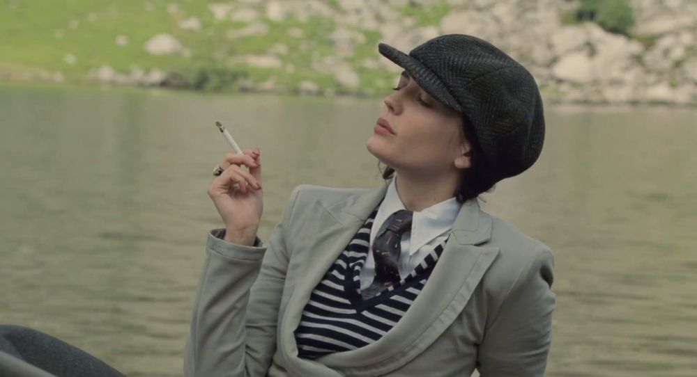 Still from the film CRACKS of Eva Green as Miss G, a white woman wearing 1930s tailored vest, jacket, tie, collared shirt and a pageboy cap, holding a cigarette and sitting with her eyes closed by a lake.