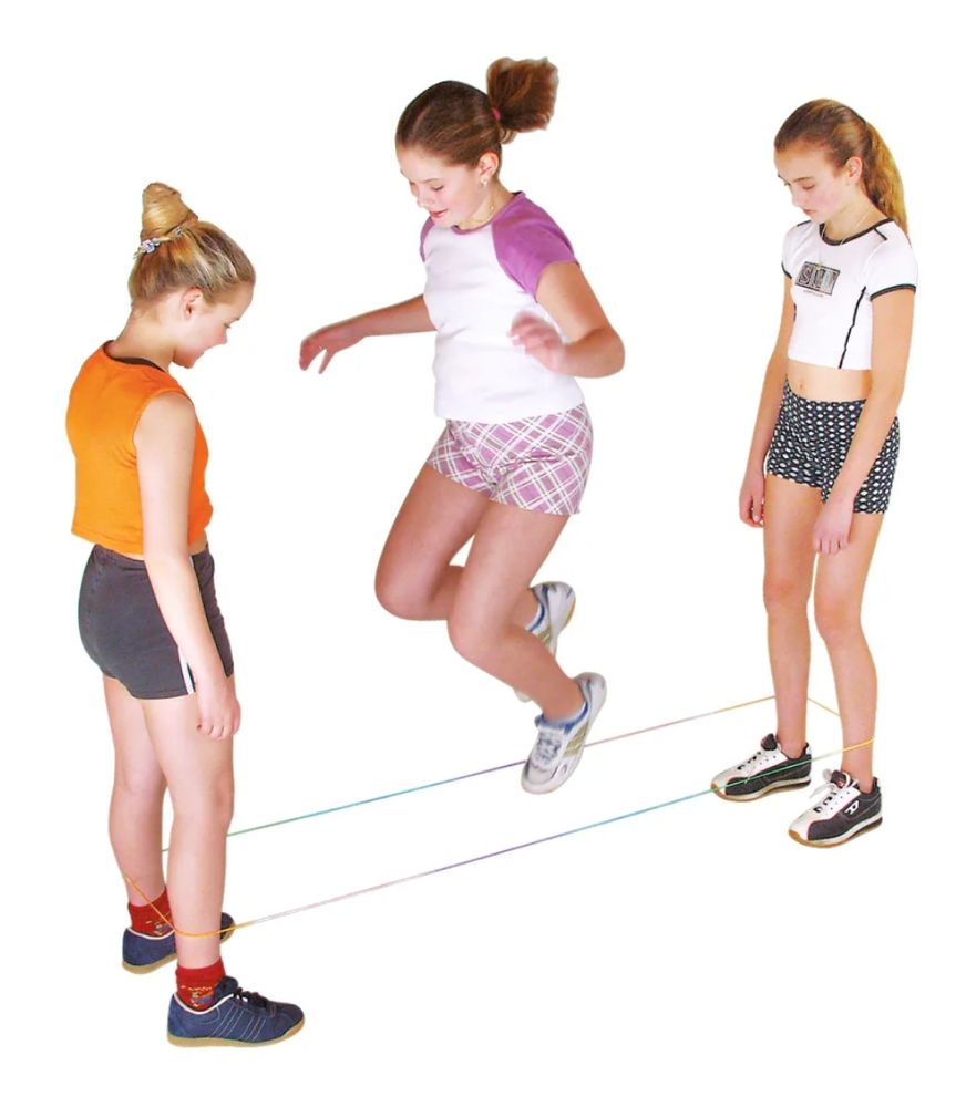 3 Mädchen spielen Gummi-Twist. 2 stehen außen und spannen etwas über Knöchelhöhe ein Gummiband zu einem Rechteck. Die 3. hüpft um die gespannten Gummibänder.