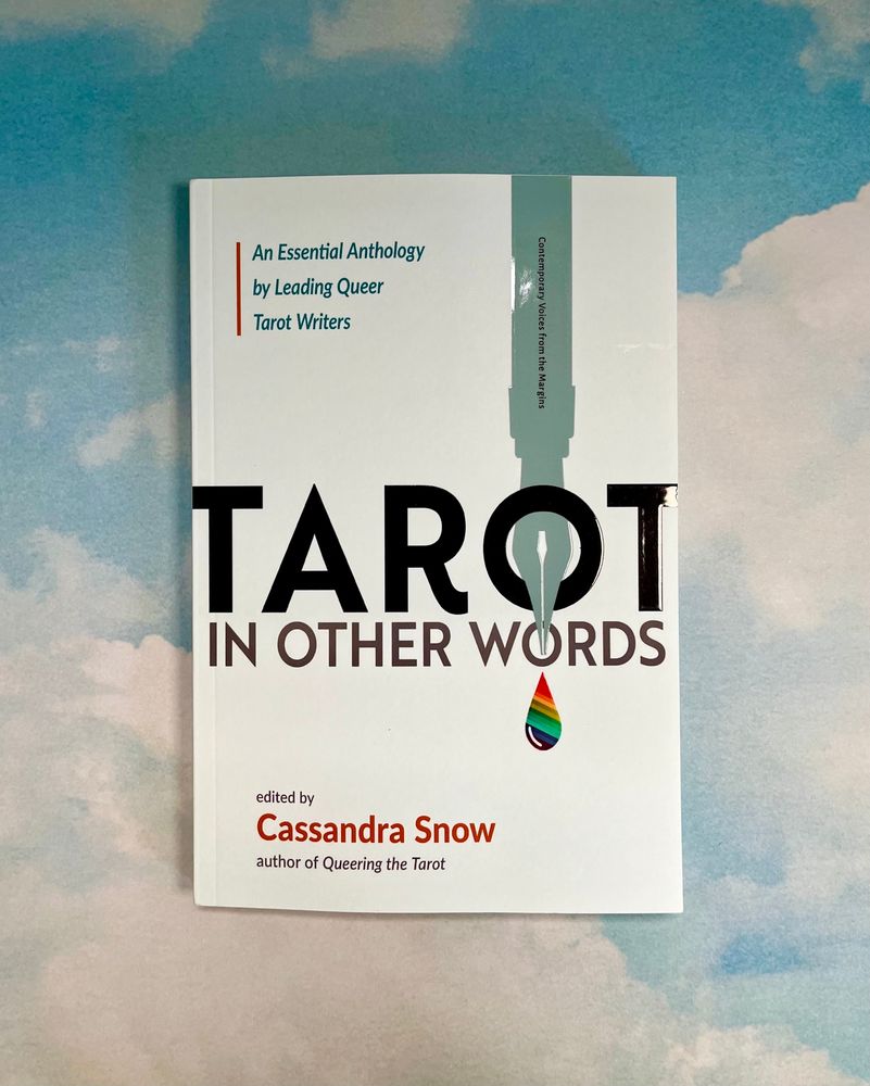 Book called “Tarot in Other Words” in photograph. The word “Tarot” in large black font on the book. A pen with rainbow ink on the white cover. Behind the book is a backdrop of a light blue sky with fluffy clouds.