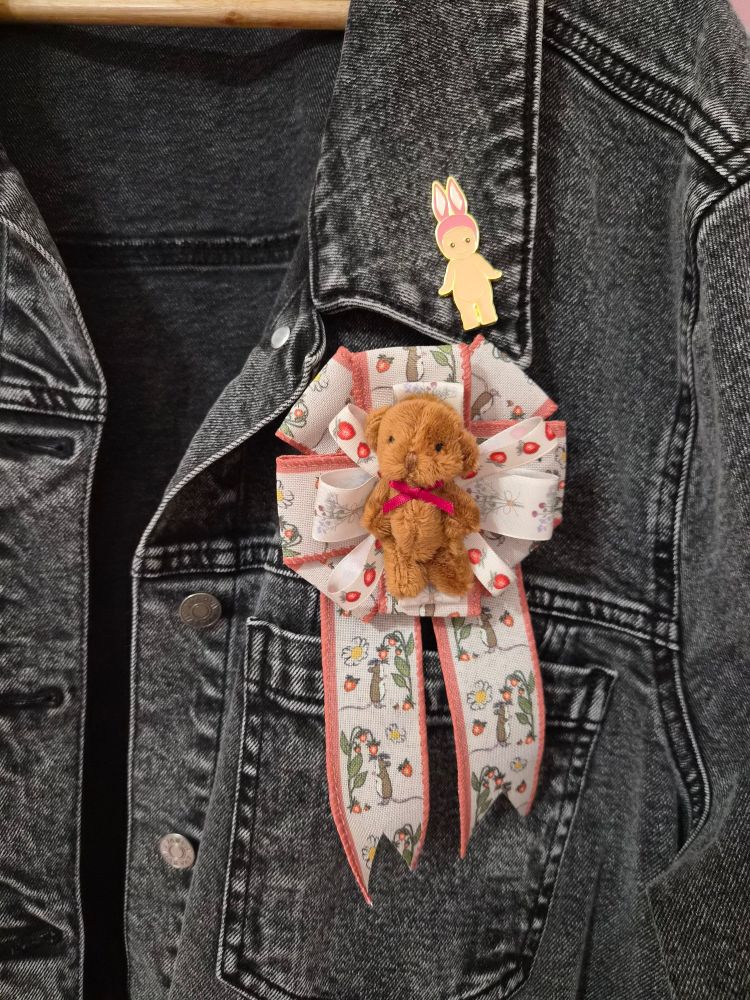 A prize ribbon with a miniature brown teddy bear as the centerpiece, pinned to a denim jacket.