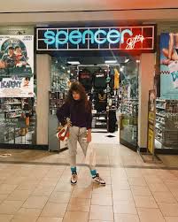A woman from the 80s with big 80s hair standing in front of a Spencer Gifts store in a mall. 
