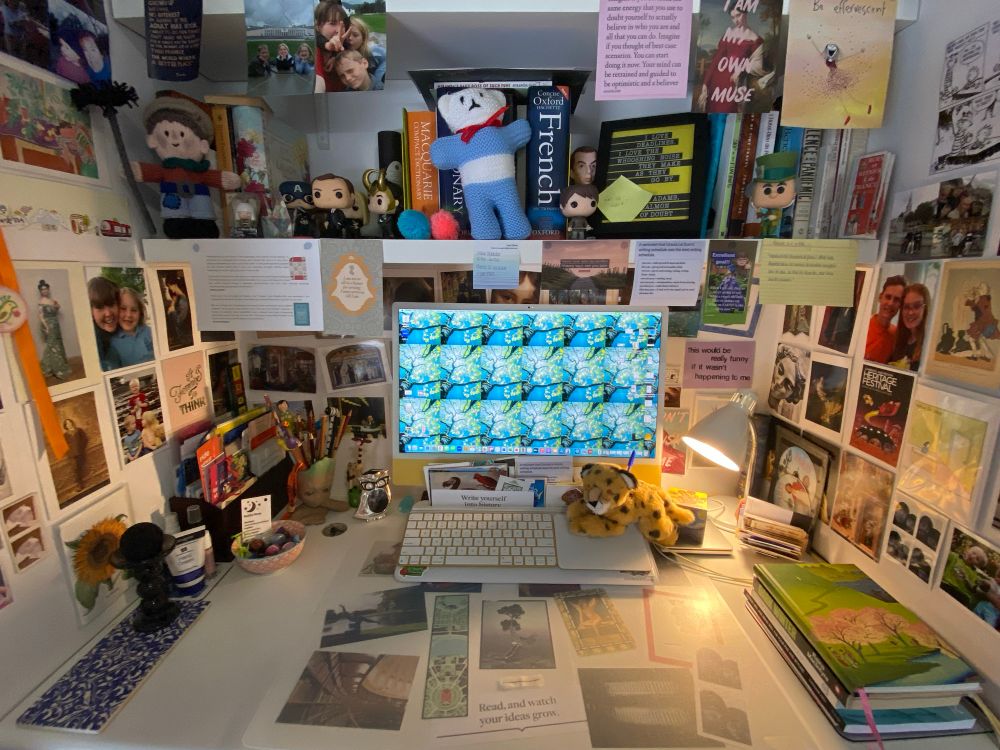 Photograph of my desk featuring in central position my yellow iMac upon which is a tiled desktop photograph of a tree photographed from the bottom looking up at the gorgeous canopy of light green almost luminous leaves and a gorgeous blue sky. Surrounding that are a heap of photographs and postcards of Family and thesis topic and places I’ve been because my desk isn’t window facing. There are also a lot of pop figurines and a knitted chimney sweep for luck from my Nanna and a blue and white knitted teddy that I got when I was in hospital two years ago from the fundraising gift shop in the foyer. You can also see the first row of bookshelves with reference books and dictionaries and a little stack of thesis books as well.