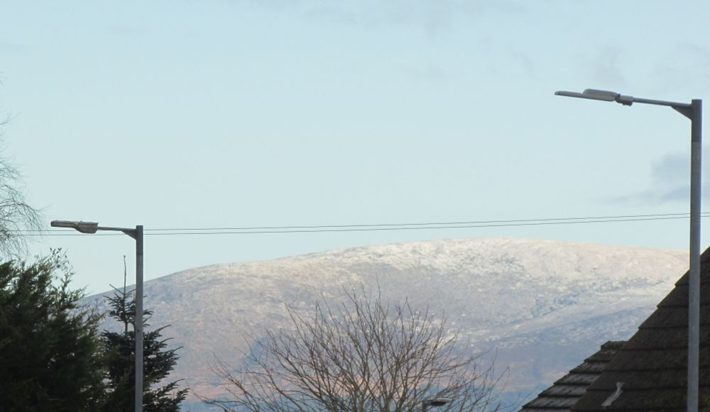 A photo of part of the Galloway Hills with a dusting of snow on the top.
In the foreground are a couple of lamposts, a few trees and the edge of a couple of roofs.