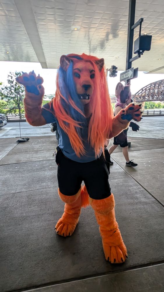 An orange anthropomorphic lion, wearing black shorts, a blue shirt.  They have a long orange mane with blue stripes.  Standing in front of a street, roaring at the camera