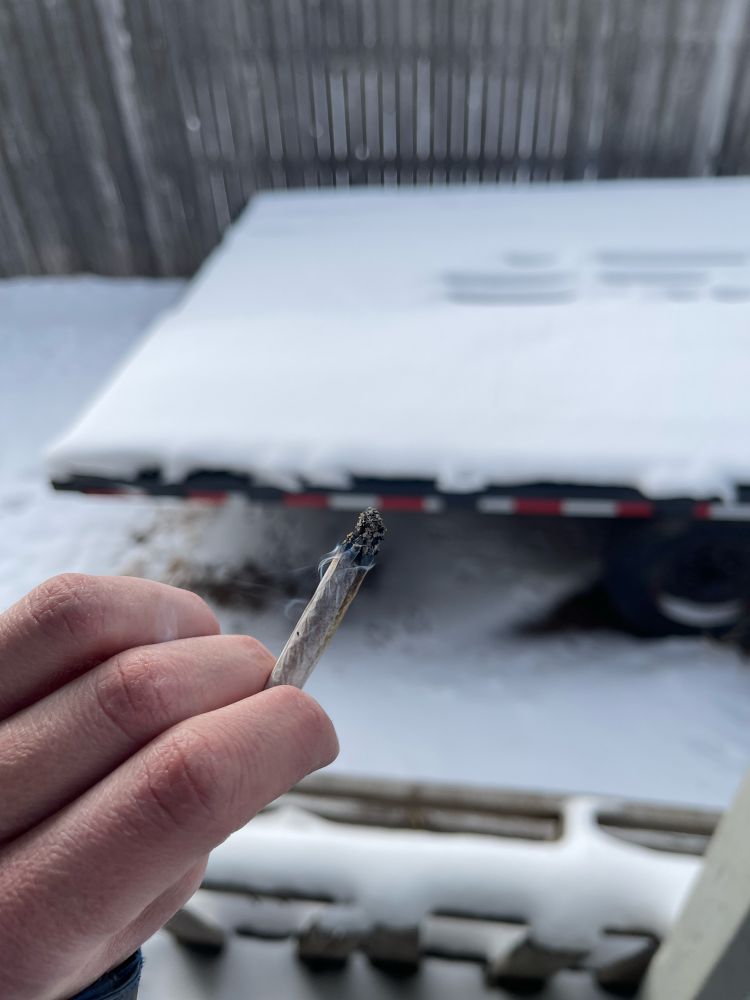 A close-up of a person’s hand holding a lit joint with ash on the tip and a wisp of smoke. The background shows a snow-covered flatbed trailer with faint markings in the snow, a wooden fence, and a winter setting.