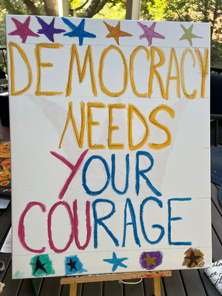 sign on an easel reading “DEMOCRACY NEEDS YOUR COURAGE” with “OUR RAGE” highlighted in blue