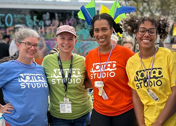 Four smiling people at an outdoor festival standing side by side wearing colorful shirts with logos on them that say NOMADstudio. In the background is a bus that is being colorfully painted by people in attendance.