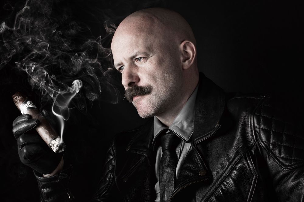 Male, shaved head, moustache. Looking to the left. Wearing a grey leather shirt and black leather tie, black biker jacket on top a JFR Lunatic smoulders in his gloved hand.