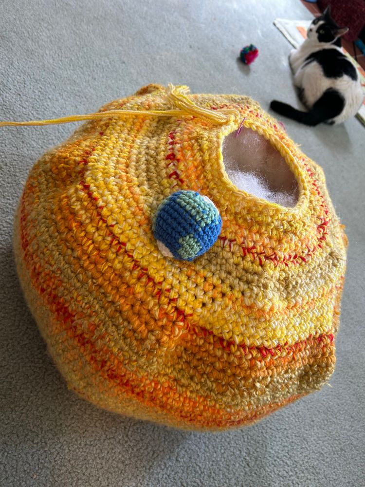 A tiny crocheted Earth sits on an unfinished crocheted Sun. A tuxedo cat sits in the background unconcerned with the celestial wonder evolving in yarn and synthetic stuffing. 