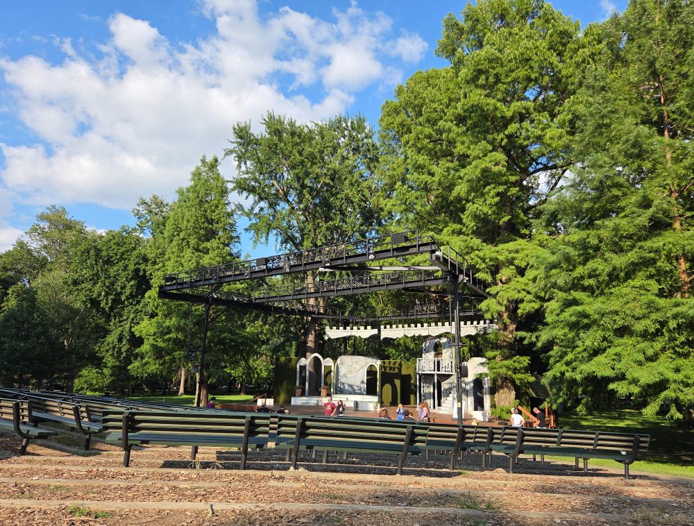 A castle-like stage design for Twelfth Night beneath the towering trees of Central Park, a blue sky, and fluffy white clouds.