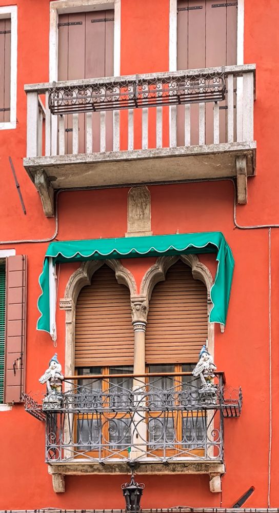 Übereinander jeweils zwei Fenster nebeneinander in einer orangefarbenen Hausfassade. Die unteren beiden Fenster haben ein schön gestaltetes Metallgitter davor und eine grüne eingezogene Markise drüber. Die Rollos sind halb runter gelassen. 
Die oberen beiden Fenster haben geschlossene Fensterläden und einen kleinen steinernen Balkon davor.