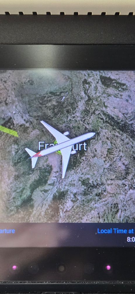 A close-up of an in-flight entertainment screen shows a digital map with a plane graphic hovering over Frankfurt, Germany, indicating the aircraft’s current location. The terrain below is depicted in satellite imagery, and part of the user interface shows the local time in the corner.