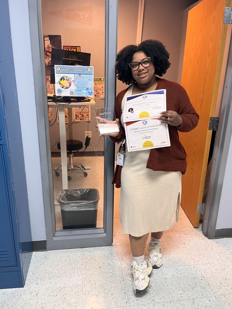 Murissa Mays standing proudly in front of her office, holding two certificates and a clear glass award. One certificate reads “Visionary for Equitable Technology Implementation” and the other recognizes Frontier Schools for its “STEAM Culture of Innovation.” A personalized office poster with her name is visible on the window behind her.
