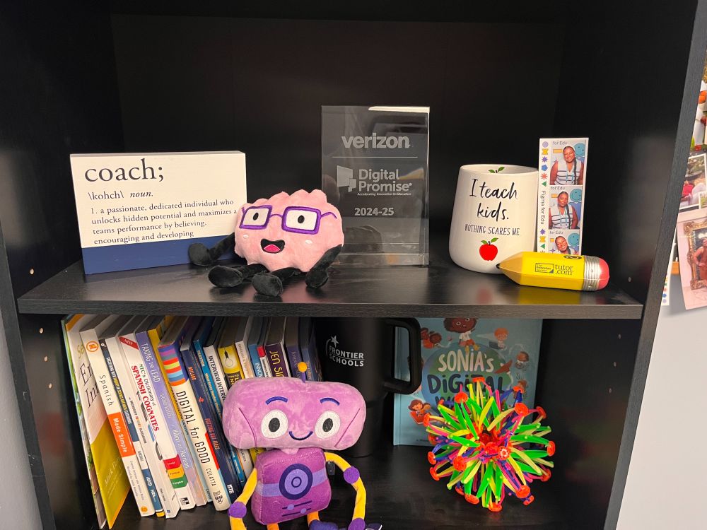 educational items. The top shelf displays a plaque for Digital Promise’s Verizon Innovative Learning Schools 2024-25 award, a mug reading “I teach kids. Nothing scares me,” a “coach” definition sign, a plush brain with glasses, and a photo booth strip. The bottom shelf contains books, a Frontier Schools mug, a colorful Hoberman sphere, a plush purple robot, and a children's book titled Sonia's Digital World.