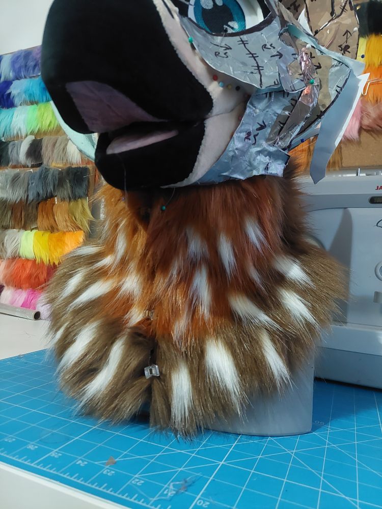 Photo of a bird fursuit's neck - it is rust and chocolate coloured with 26 white specks throughout the front of the neck. The beak is black with a pink inner mouth, and one teal eye is visible looking down at the camera.