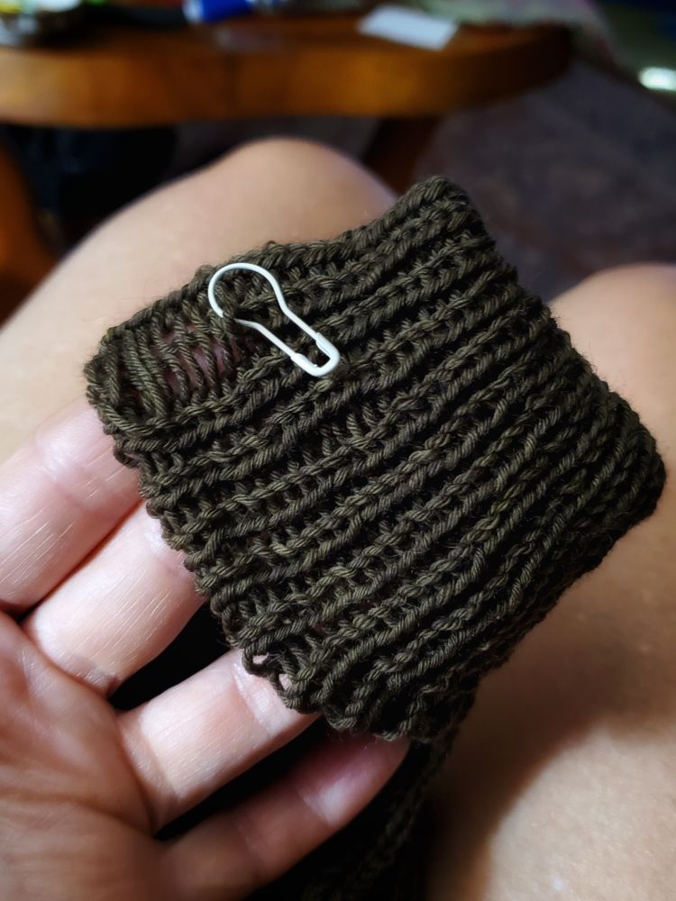greenish knitted cuff stretched over fingers to show dropped stitch captured on a white bulb pin.  #knitting 🧶 #knitsky