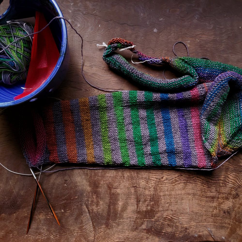 partial knitting sleeve of gray self striping roy g biv, heavy on the greens on a wood burl table
