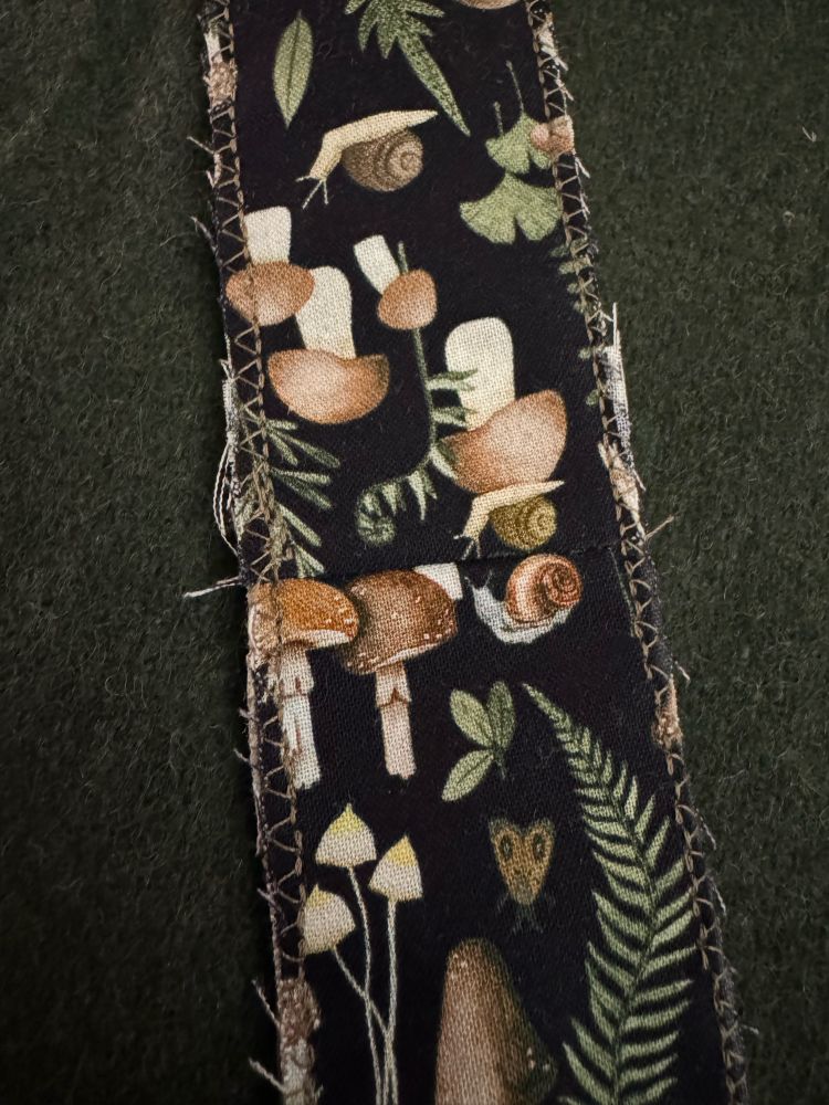 second strap; same description: A shoulder strap in progress, made with a black-background mushrooms, ferns, and snails-printed cotton fabric piece; it is long and rectangular and the edges have been very sloppily zig-zag stitched, but where the shoulder seam of the pattern piece meets, the fabric is pieced together so that the directional fabric print will be upright from both front and back of the strap.