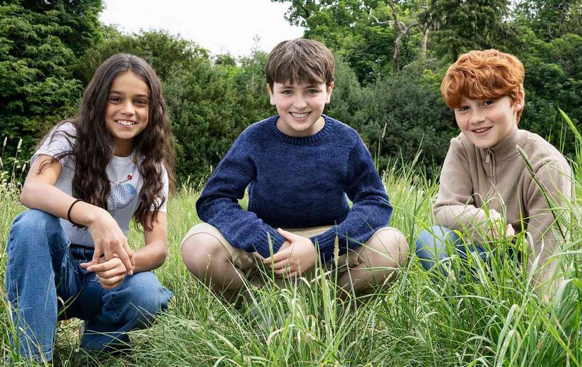 Foto de três crianças agachadas em um gramado. Uma menina de pele morena clara e cabelo castanho ondulado comprido, um menino de pele branca e cabelo castanho curto e um menino de pele branca e cabelo ruivo curto. Os três são os protagonistas da nova série de Harry Potter.