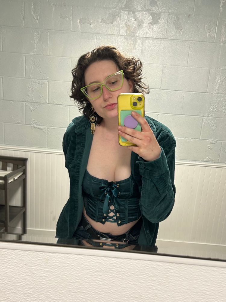 Mirror selfie of me, a white enby person, wearing a teal corset top under a matching teal button-up corduroy shirt, green cat eye frames, and brass dangle earrings. The shirt is open and I am slightly leaning towards the mirror. 
