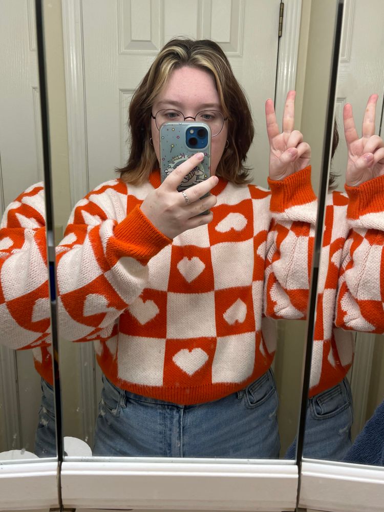 Person with brown and blonde hair in orange and white checkered heart sweater and blue jeans. They’re taking a mirror selfie and holding up their blue iPhone in one hand and a peace sign in the other 