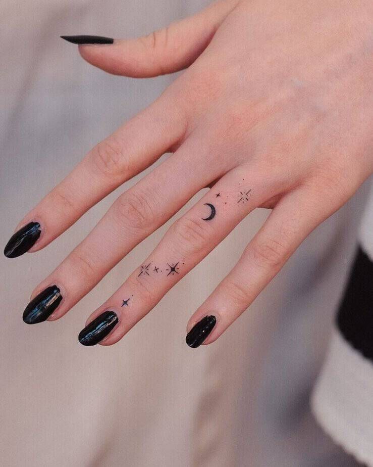 A white person’s hand outstretched with black nail polish and on their ring finger tattoos of a moon and stars 