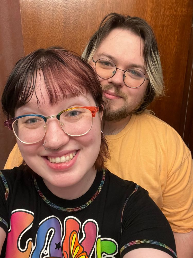 In the front op stands in a black and rainbow shirt that says love. They have split dye black and purple hair with rainbow glasses. Their hair is pulled back into a half pony tail. Behind her stands a tall handsome man with brown roots overtaking his blond tips. He had an orange shirt on and round black glasses. Both are smiling happily. He also has a really cute mustache and beard 
