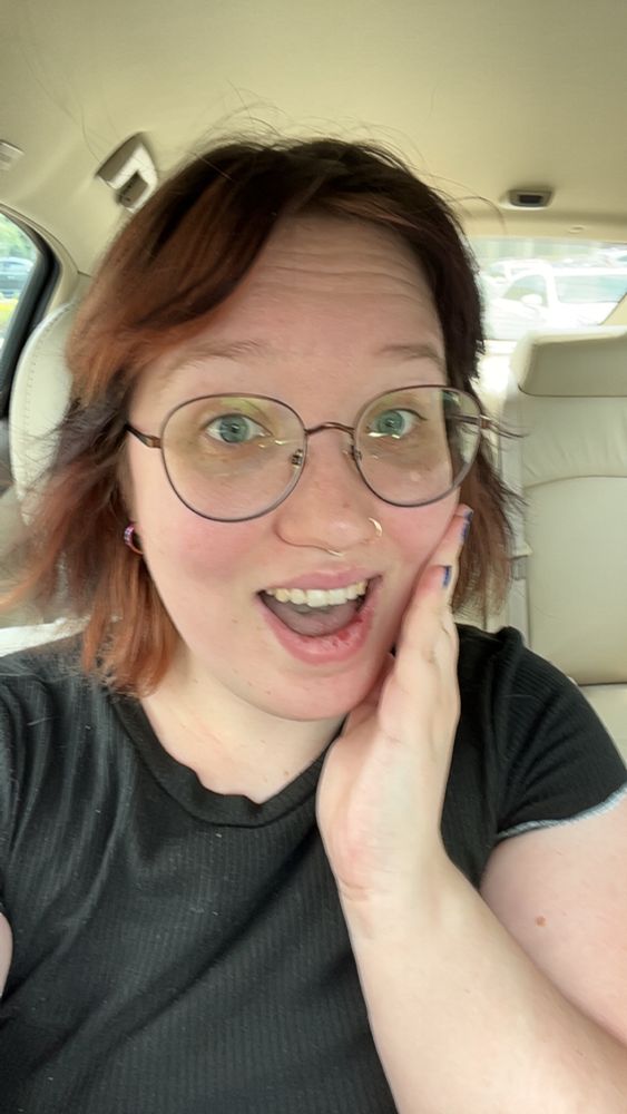 OP with black hair and reddish highlights smiling with her hand on her face showing off round metal brown glasses