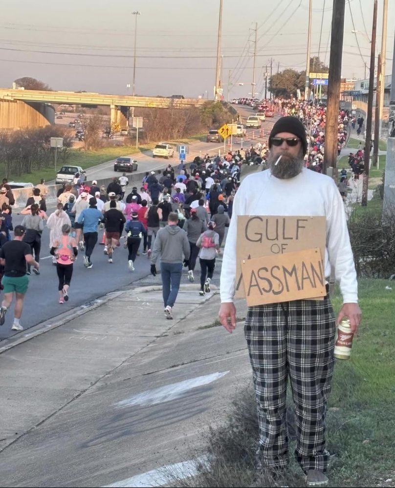 A man holding a very large Miller High Life can stands alongside the route of the Austin marathon wearing a cardboard sign that reads Gulf of AssMan.