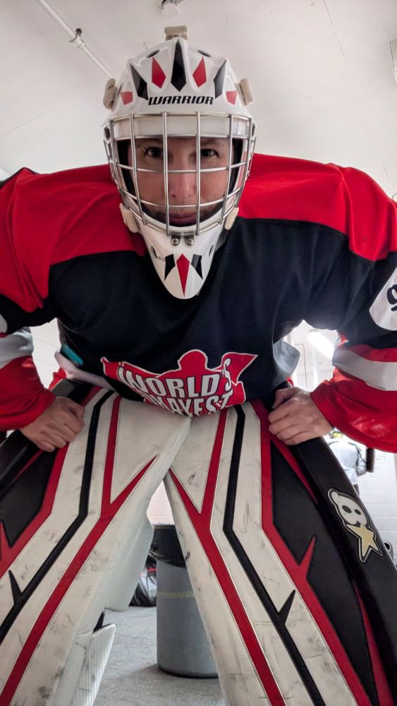A hockey goalie in red, black, and white leans forward and stares into the camera with un-gloved hands on the thigh rises of her pads
