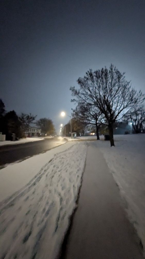 Street lights in the distance illuminate a winter scene - a shiny wet road borders a snow-covered field with a recently plowed sidewalk stretching away from the camera. The branches of a tree with few leaves left make dark scribbles against the sky, where low cloud reflects the city's glow