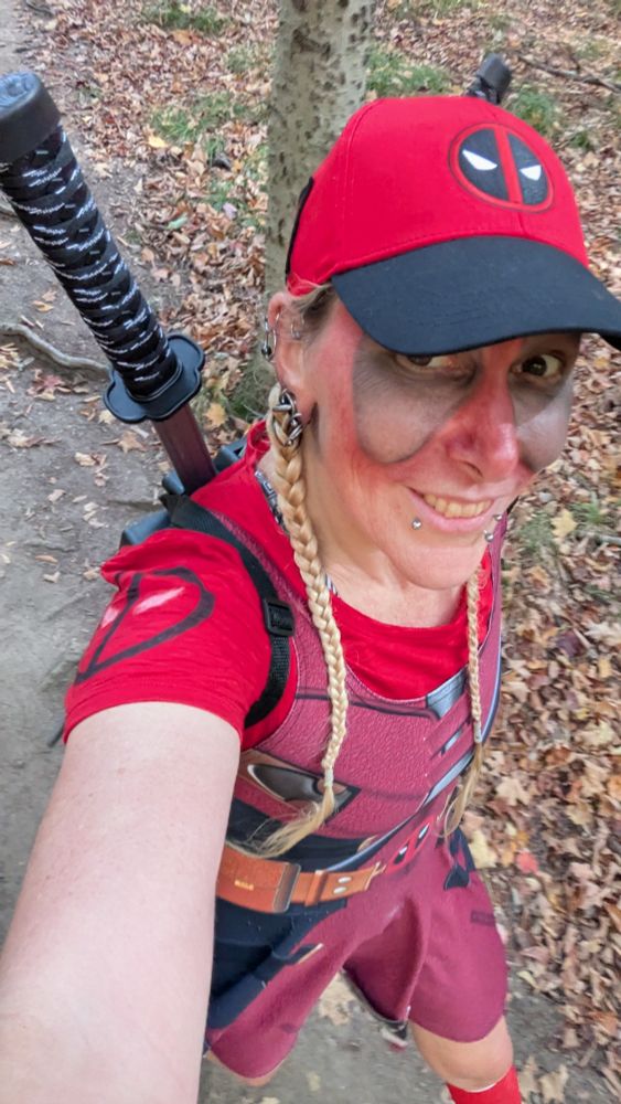 A woman with two blonde braids wears a red cap with a Deadpool logo and black brim, a red shirt with a Deadpool logo on the shoulder, a dress that bears a remarkable resemblance to a Marvel cartoon character, twin katana swords crossed on her back, and red and black face paint. She is moving on foot along a dirt path with exposed roots, bordered by drifts of fallen leaves