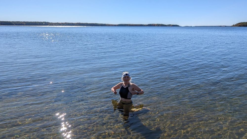 A woman in a toque and bikini kneels in the crystal clear sunlit waters of Georgian Bay, giving thumbs up with both hands