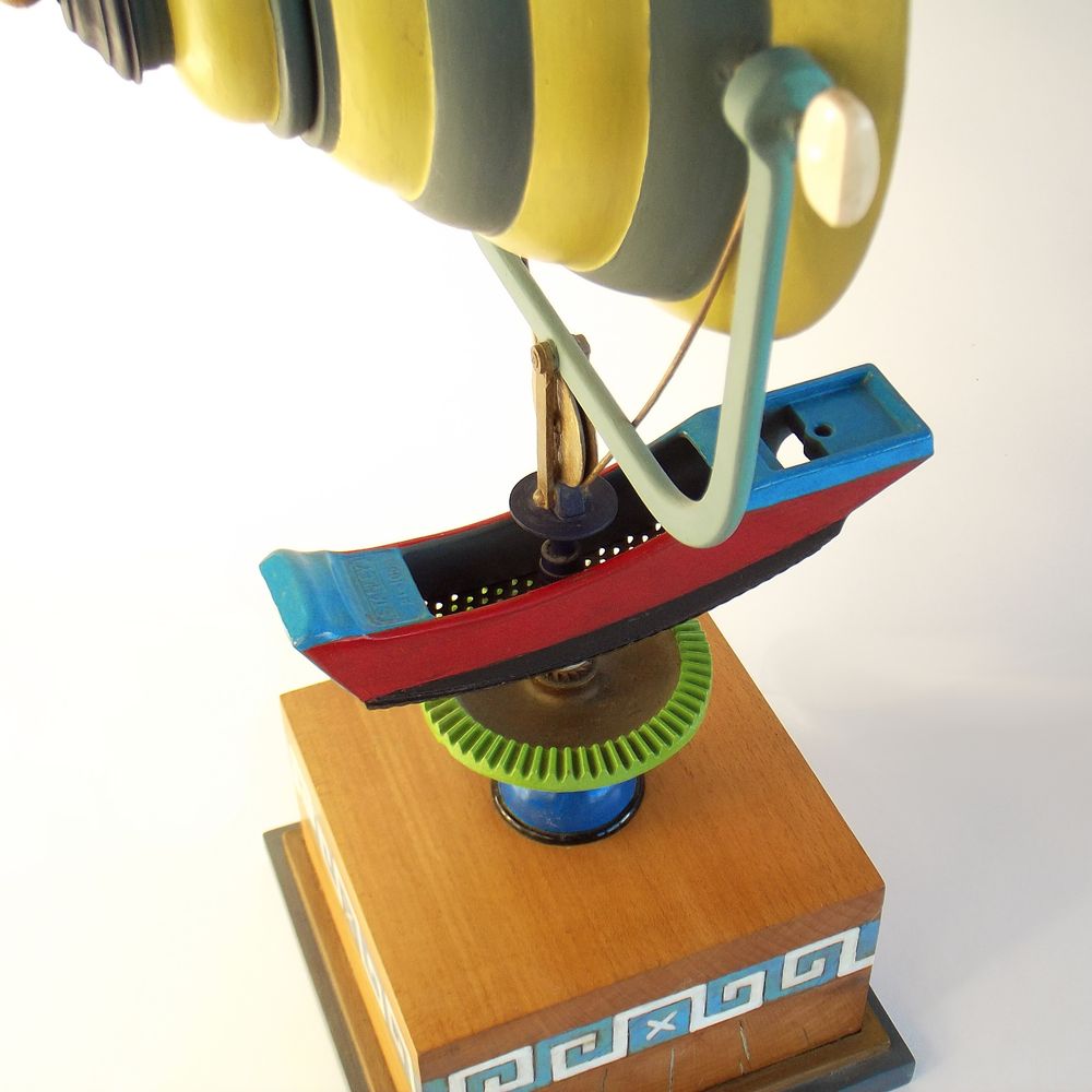 Close-up detail photograph of an abstract assemblage sculpture titled Muse ΜΕΛΕΤΗΝ by Charlayn von Solms. Visible is a carpenter's rasp mounted on a hand-drill gear and a wooden plinth painted with a Greek key pattern.