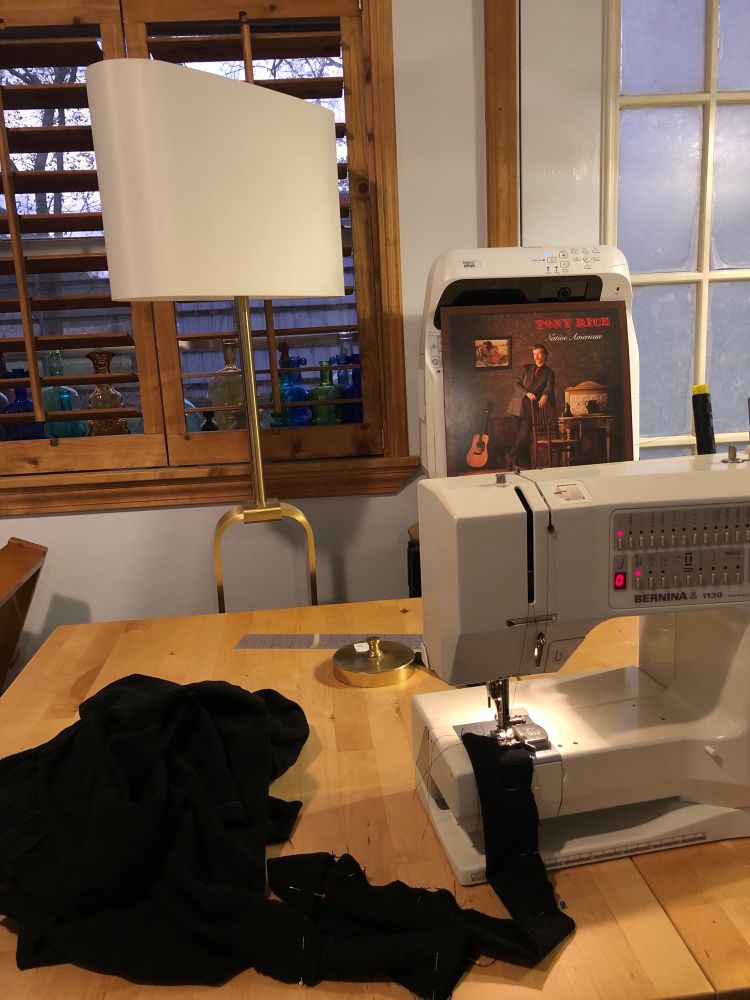 Bunched up black fabric with ties being sewn into a skirt with a Bernina sewing machine sitting on a wood table. There is a window in the background with colorful glass fiddle bottles, a floor lamp, and a Tony Rice “Native American” vinyl record sleeve leaning against a projector. 