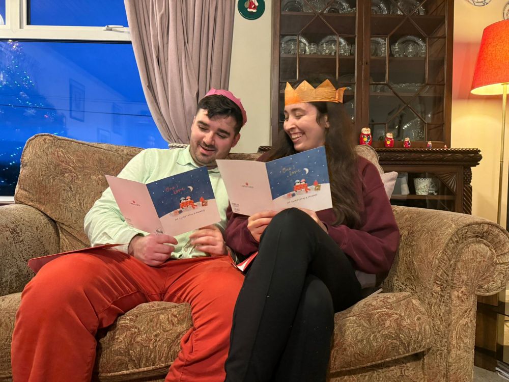 Charles and Jen sitting side by side on a sofa, wearing Christmas cracker hats and reading their identical cards