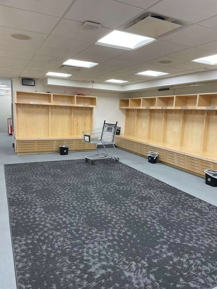 Another view of the locker room showing multiple wooden locker stalls, a shopping cart in the center of the dark carpeted area, and bright ceiling lighting. The clean, organized space features a neutral color scheme and practical layout.