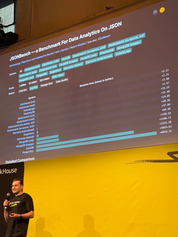 A man smiles holding a microphone beneath a large slide on Json performance where clickhouse is very performant and Postgres and DuckDB are least 