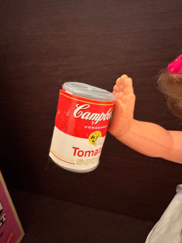 A close up of a miniature can of Campbells soup magnetically attached to the flat of a doll’s hand
