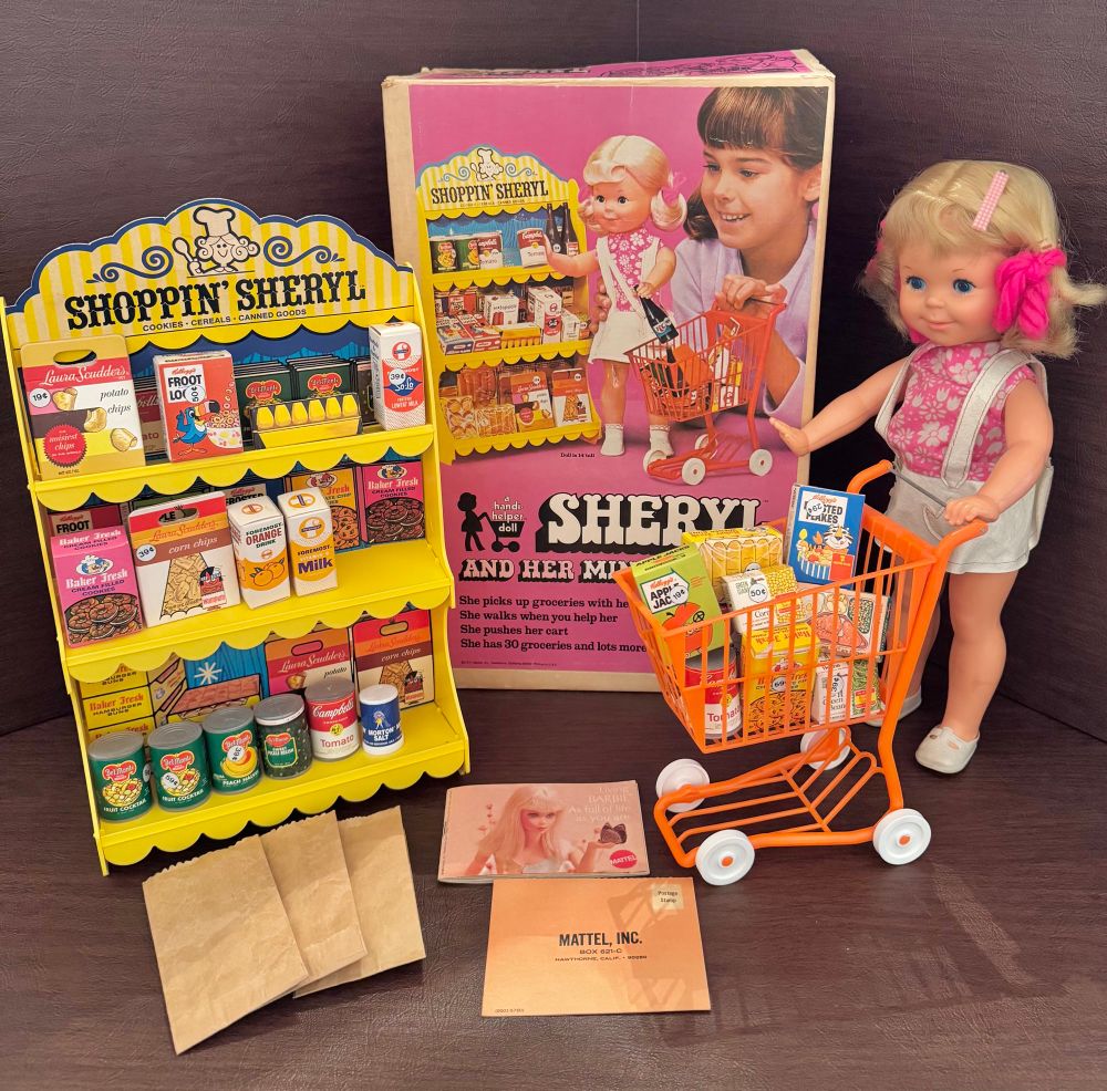 A blonde juvenile doll pushing an orange shopping cart full of grocery boxes beside a market playset with a large box in the background that reads Sheryl and her mini market 