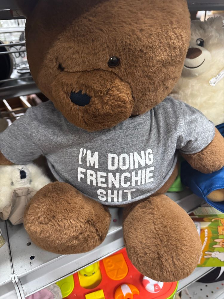 A stuffed dark brown toy teddy bear sitting on a crowded metal store shelf, wearing a grey shirt with white printing that reads I’m doing frenchie shit 