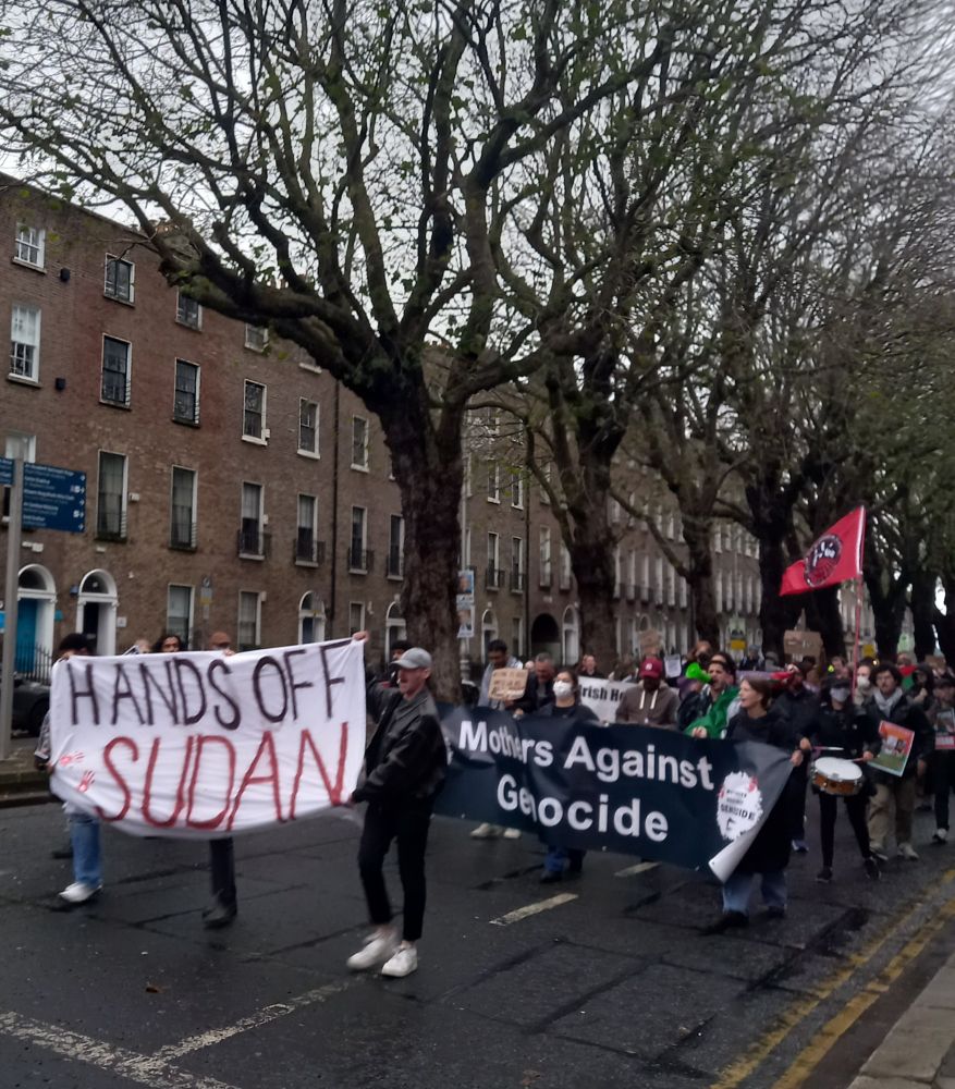 Dublin, group of people walking in the middle of the road with red bricks building and leafless trees in the background. The protests are carrying flags and homemade signs, in the front a banner "Mothers Against Genocide" and a white sheet with painted words "HANDS OFF SUDAN" 
