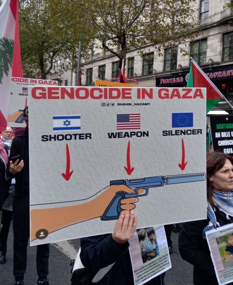 O'Connell Street, Dublin 
People walking in the middle of the street with Palestinian and Lebanese flags and signs.  The main on in the photo is a illustration of a hand holding a gun with silencer and over it 3 flags with arrows: (Israeli) SHOOTER (USA) WEAPON (EU) SILENCER